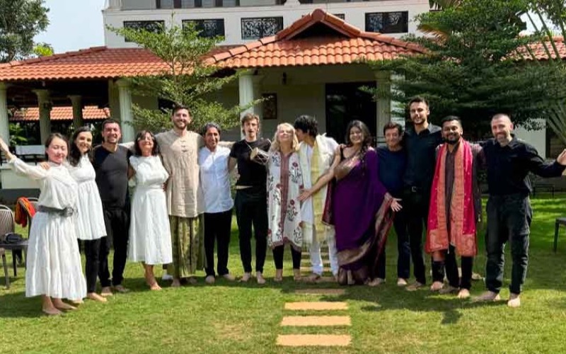 Elenco reunido no jardim de uma casa, em figurinos indianos, ao fim de uma montagem.