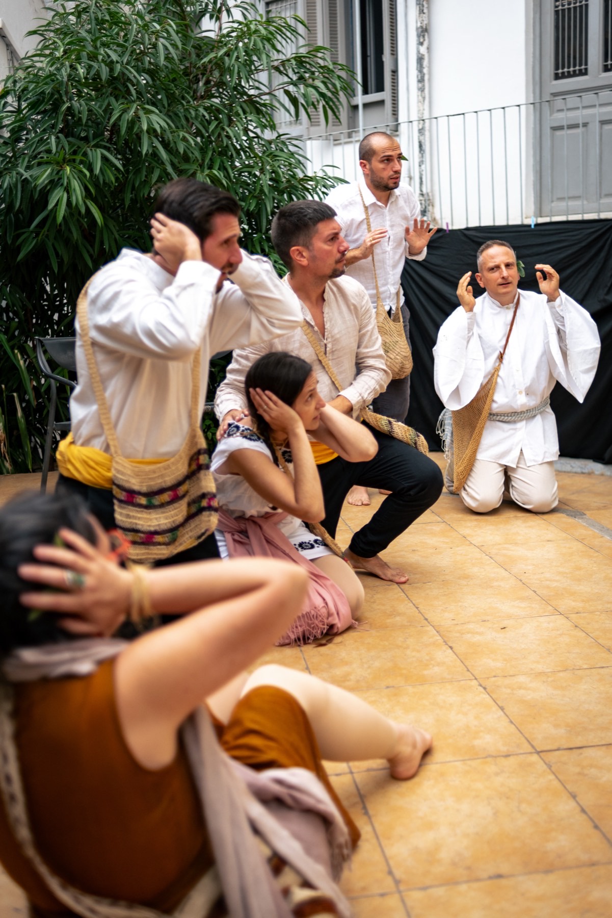 Coro de atores ajoelhados, mãos sobre os ouvidos, em cena.